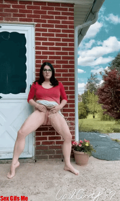 A girl with glasses leans against a wall so that she can piss standing up.